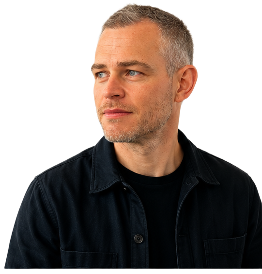 Tom Sharp, wearing a dark overshirt over a black t-shirt, turned slightly towards the page.